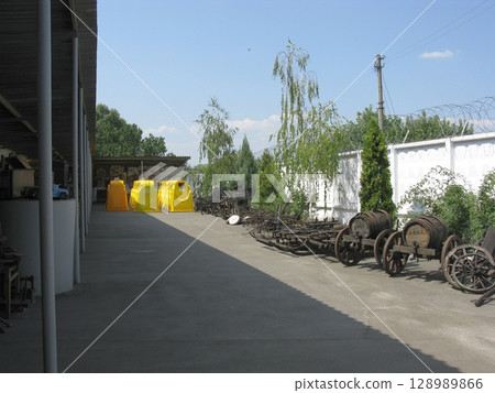 An outdoor display of weathered wooden carts and barrels near a white fence with trees under a clear blue sky An outdoor display of weathered wooden carts and barrels near a white fence with trees under a clear blue sky 128989866