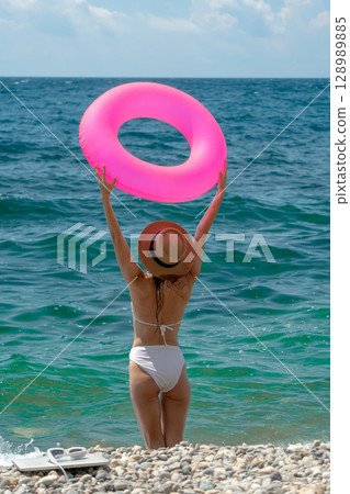 Woman Beach Sea. Young woman in white bikini holds pink ring, ready for summer swim in blue sea. 128989885