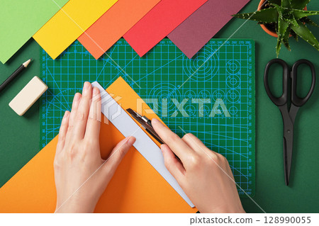 Hands cutting colored paper with utility knife on green cutting mat in top view. Creative craft process, handmade project preparation. 128990055