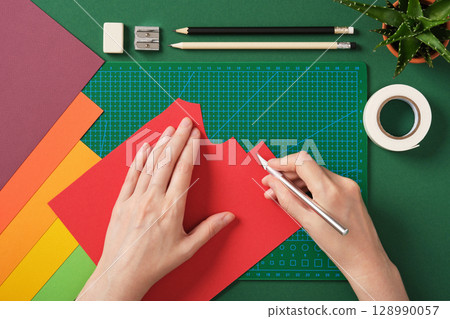 Hands cutting red paper on cutting mat with stationery around. Idea of craft, handmade decoration and creative paper DIY design project. Hands cutting red paper on cutting mat with stationery around. Idea of craft, handmade decoration and creative paper DIY design project. 128990057