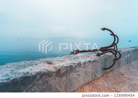 A rusty anchor is sitting on a concrete wall next to the ocean 128990484