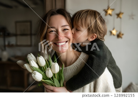 A happy mother embraces her child receiving a sweet kiss, celebrating this beautiful moment with a fresh white tulip bouquet. 128992155