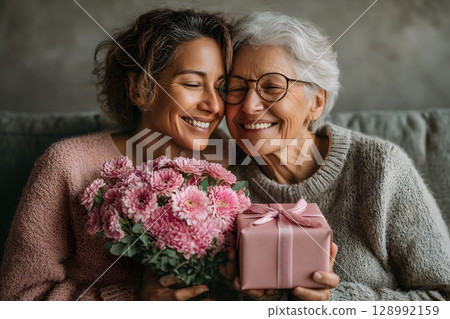 A loving mother and her happy daughter share a warm embrace, holding a vibrant pink flower bouquet and a thoughtful gift. 128992159