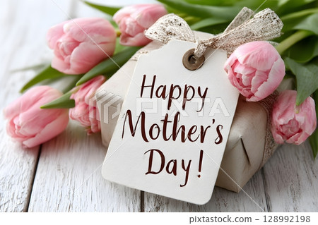 A festive gift box displaying a Happy Mother's Day tag rests among vibrant pink tulips on a rustic wooden table. 128992198