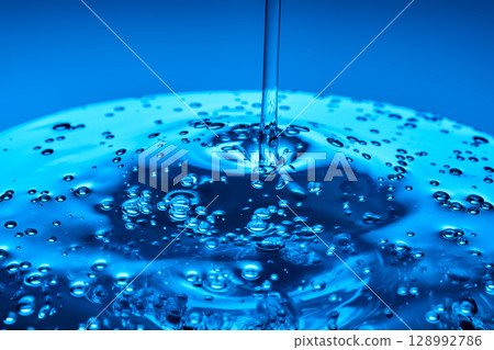 The viscous gel serum for the face flows and collects into a drop on a blue background.. The viscous gel serum for the face flows and collects into a drop on a blue background.. 128992786