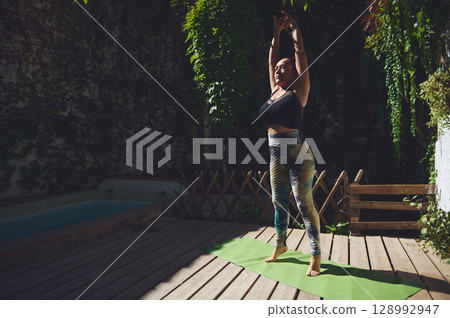 Woman Performing Yoga Outdoors on a Wooden Deck in a Serene Garden Setting Woman Performing Yoga Outdoors on a Wooden Deck in a Serene Garden Setting 128992947