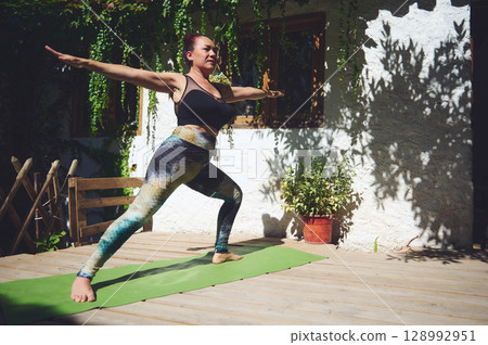 Woman Practicing Yoga on Wooden Deck Outdoors in Warrior Pose Woman Practicing Yoga on Wooden Deck Outdoors in Warrior Pose 128992951