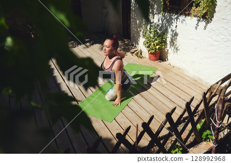 Woman Performing Yoga on Outdoor Deck Surrounded by Natural Light and Greenery Woman Performing Yoga on Outdoor Deck Surrounded by Natural Light and Greenery 128992968