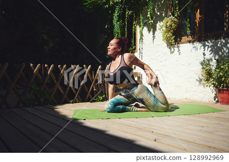 Woman Practicing Yoga Outdoors on a Green Mat in a Sunlit Patio with Lush Foliage Woman Practicing Yoga Outdoors on a Green Mat in a Sunlit Patio with Lush Foliage 128992969