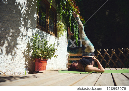 Woman Practicing Shoulder Stand Yoga Pose Outdoors on a Wooden Deck Woman Practicing Shoulder Stand Yoga Pose Outdoors on a Wooden Deck 128992976