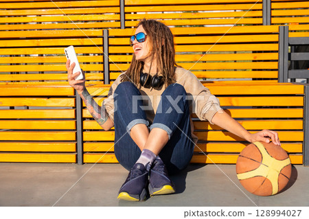 Woman with dreadlocks and sunglasses sitting next to a basketball, holding phone and looking sideways. Woman with dreadlocks and sunglasses sitting next to a basketball, holding phone and looking sideways. 128994027