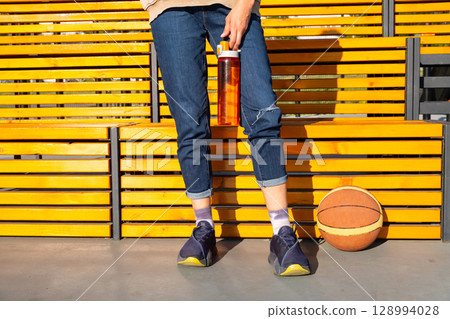 Woman standing with a water bottle and a basketball next to a yellow wooden bench. 128994028