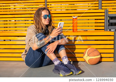 Woman sitting on basketball court, using smartphone with headphones. 128994030
