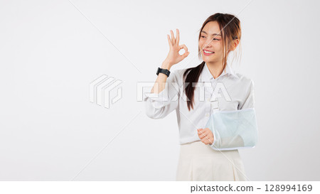 Happy young asian woman injury arm broken with a soft splint and sling makes an ok gesture after her accident showing she is doing fine and recovering well from her accident, isolated white background Happy young asian woman injury arm broken with a soft splint and sling makes an ok gesture after her accident showing she is doing fine and recovering well from her accident, isolated white background 128994169