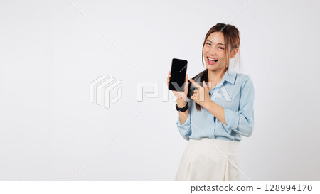 Excited Asian woman holding a mobile phone, pointing a finger to the blank screen for showing. She is happy with her smartphone in hand. isolated studio white background 128994170