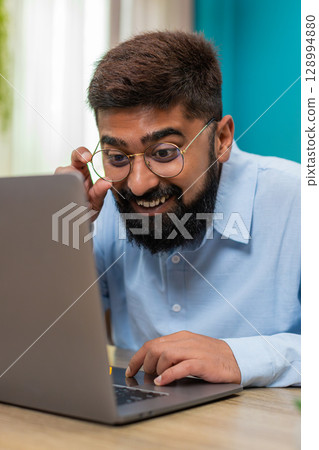Indian man smiles after email reply on laptop nods celebrating project finish success indoors table Indian man smiles after email reply on laptop nods celebrating project finish success indoors table 128994880