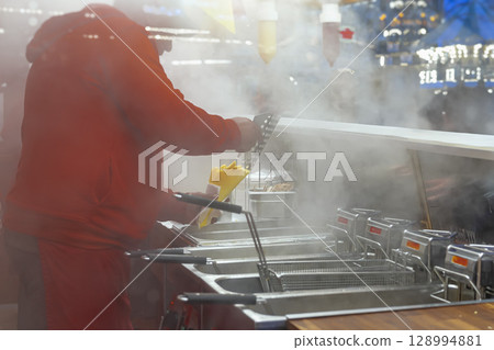 Cook wearing red prepares steaming batch of fresh French fries at busy outdoor food stall. Concept of fast food, street cooking, hot winter snacks Cook wearing red prepares steaming batch of fresh French fries at busy outdoor food stall. Concept of fast food, street cooking, hot winter snacks 128994881