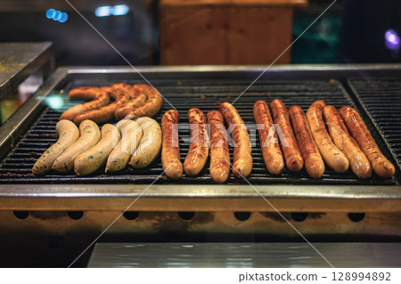 Grilled sausages. Variety of grilled sausages sizzling on barbecue at food market with golden crusts. Concept of sausages, street food, festive grilling 128994892