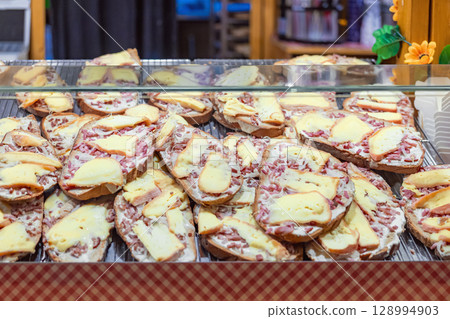 Rustic bread slices topped with melted cheese and bacon displayed at festive food stall. Concept of comfort food, hearty snack, winter street flavors Rustic bread slices topped with melted cheese and bacon displayed at festive food stall. Concept of comfort food, hearty snack, winter street flavors 128994903
