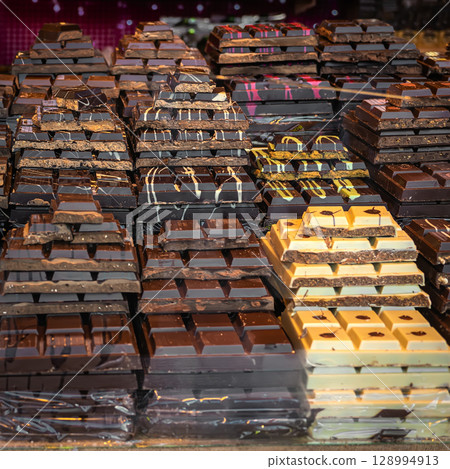 Chocolate bars in confectionery shop display case. Stacks of assorted chocolate bars in various flavors and colors arranged at market stall. Concept of cocoa products, sweet indulgence, gourmet treats 128994913