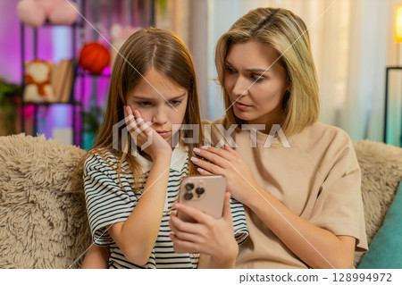 Child girl kid playing mobile game with mother on home sofa reacting emotionally to character losing 128994972