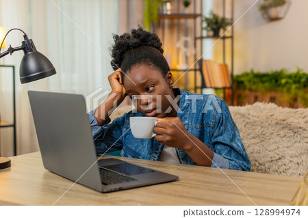 African American lazy woman drinks coffee tiredly before remote work on laptop sitting at home African American lazy woman drinks coffee tiredly before remote work on laptop sitting at home 128994974