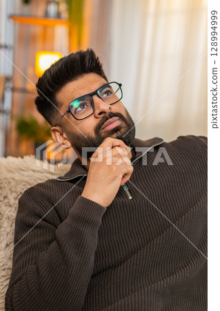 Indian young man on sofa at home looks at side with nostalgic face, thinking about someone dear 128994999