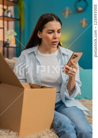 Young woman sitting on sofa unpacking delivery finding wrong item disappointed shaking head sadly 128995008