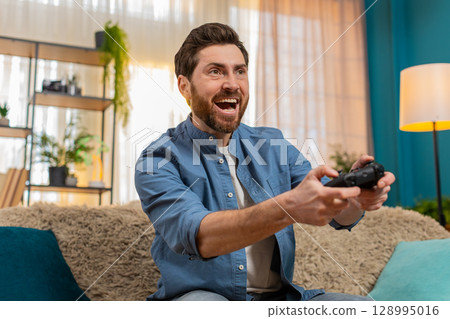 Young man intensely playing video game holding gamepad focused excited reacting home indoors sofa 128995016