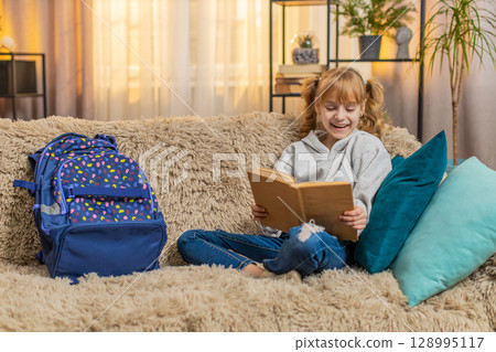 Young teenager girl reading storybook sitting at home sofa stopping at pictures gentle smile moment 128995117