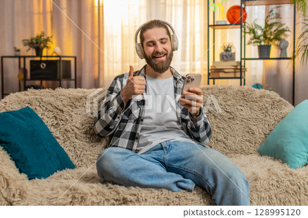 Young man in wireless headphones at home, groove, music listen, thumbs up, cool track, relaxed vibe Young man in wireless headphones at home, groove, music listen, thumbs up, cool track, relaxed vibe 128995120