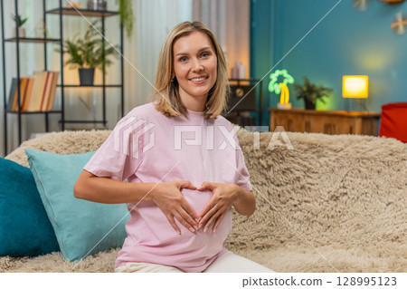 Pregnant woman forming heart with hands over belly smiling enjoying sweet loving moment before birth 128995123