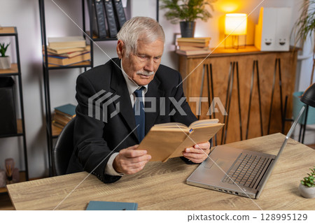 Bored Caucasian senior businessman reading book, literature, taking a rest after work at table 128995129