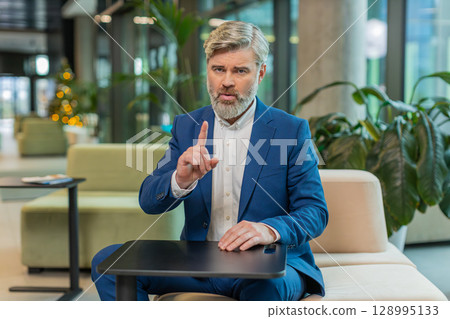 Serious mature businessman shakes finger and saying No be careful and giving advice in office lobby Serious mature businessman shakes finger and saying No be careful and giving advice in office lobby 128995133