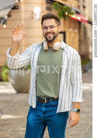 Cheerful young man smiling friendly at camera waving hands gesturing while standing on city street 128995138