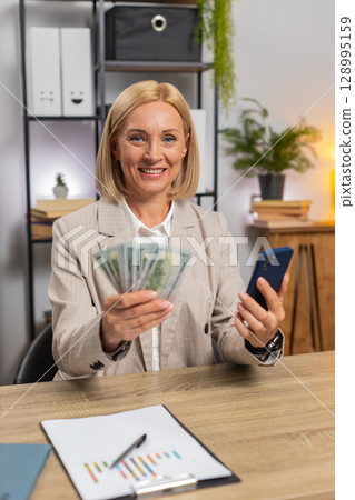 Mature businesswoman counting money dollar cash using smartphone calculator app at home office desk 128995159