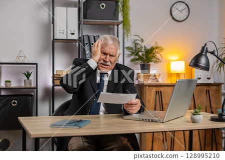 Shocked senior businessman opening envelope reading bad news in letter feels upset at office table Shocked senior businessman opening envelope reading bad news in letter feels upset at office table 128995210