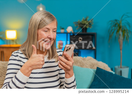 Mature woman viewing social media photos on smartphone, smiling, giving thumbs up approval at home 128995214