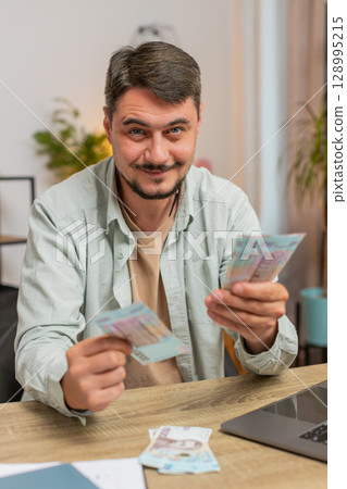 Rich happy Caucasian man counting money cash using laptop calculate domestic bills at home table Rich happy Caucasian man counting money cash using laptop calculate domestic bills at home table 128995215