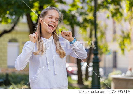 Young woman looking surprised clenching fists, shocked by sudden victory standing on city street Young woman looking surprised clenching fists, shocked by sudden victory standing on city street 128995219