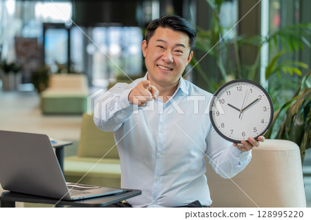 Mature businessman with laptop in lobby showing time on wall clock and pointing the finger at camera 128995220