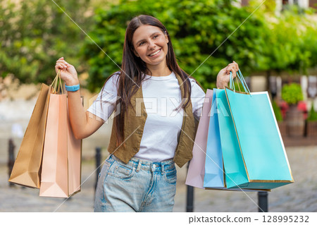 Happy smiling brunette young woman shopaholic carrying shopping bags while walking on city street 128995232