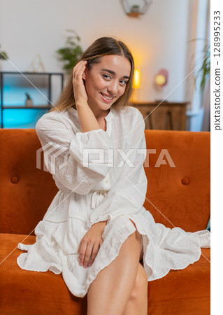 Happy attractive young woman sitting on sofa couch in living room looking shy at camera and smiling 128995233