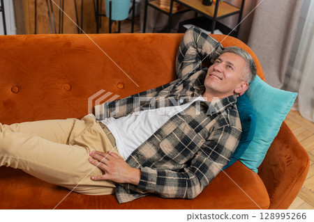 Tired young adult man sitting on comfortable couch, taking a rest daytime nap resting at home room 128995266