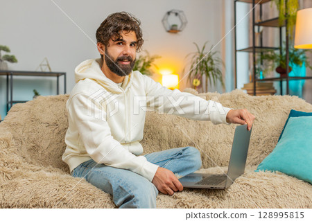 Young Indian man sitting on sofa couch, opens laptop computer start working in living room apartment Young Indian man sitting on sofa couch, opens laptop computer start working in living room apartment 128995815