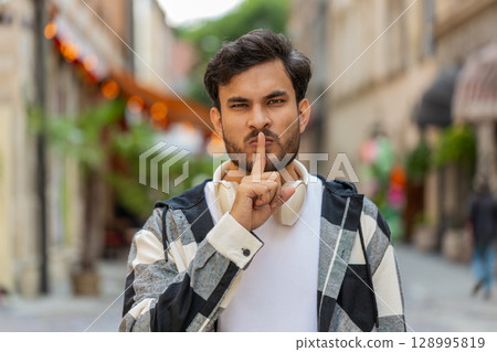 Serious Indian young man presses index finger to lips makes silence hush gesture in city street 128995819