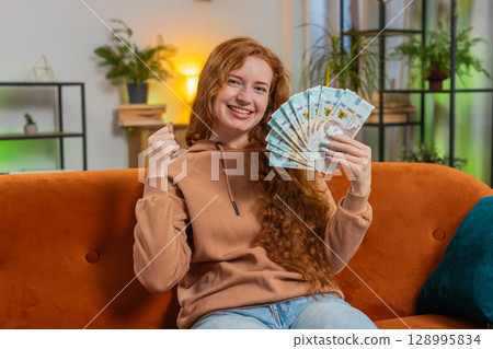 Successful rich young Caucasian woman holding euro bill money fan sitting on sofa couch at home 128995834