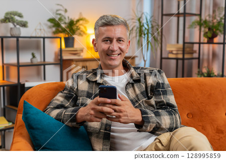 Caucasian young man sitting on couch using mobile smartphone share messages smiles at home room 128995859