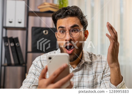 Happy amazed Indian man working on smartphone shocked by sudden victory game lottery online winning 128995883