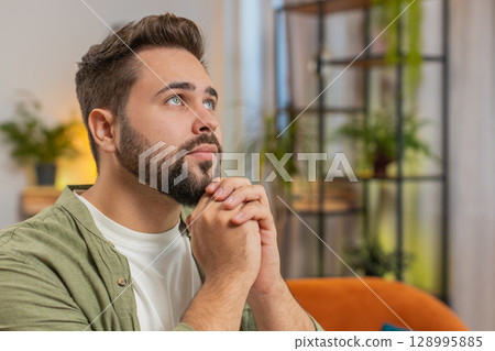 Tensed Caucasian young man sitting on sofa couch at home folding palms together pray ask for help 128995885
